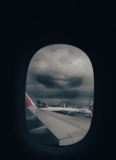 View through an airplane window showing a partly cloudy sky with dark clouds, and outside on the tarmac several aircraft parked next to each other, including one with the airline branding 'Azul'. The