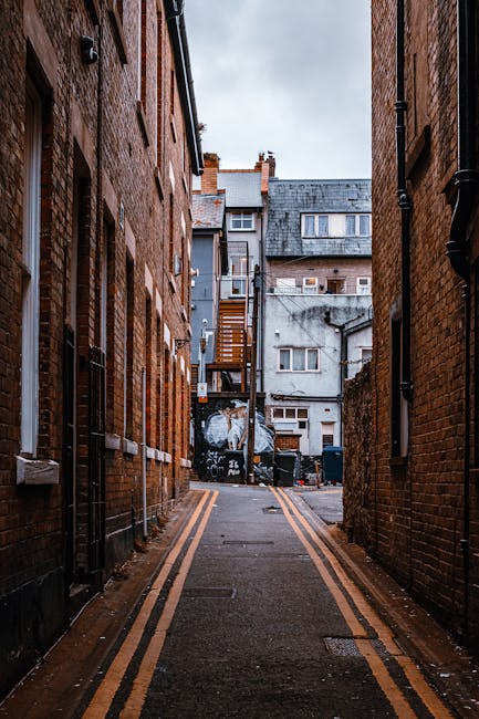 A narrow alleyway between brick residential buildings with fire escapes and scaffolding visible on the upper floors, leading to a broader street in the background. The alley is dimly lit with shadows
