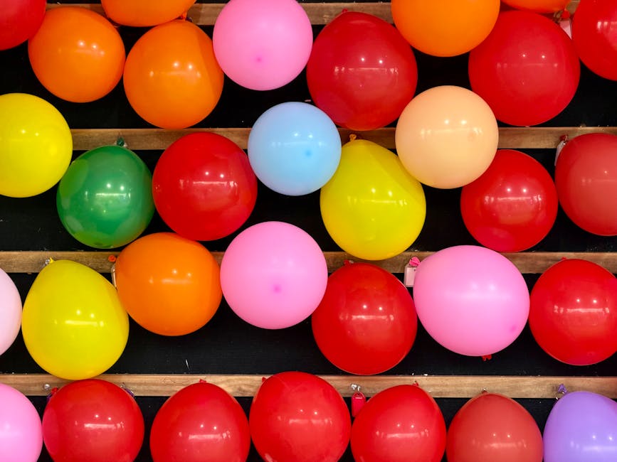 A close-up view of numerous colorful balloons, including pink, red, orange, yellow, green, and light blue, arranged in rows on a black surface with wooden slats. The balloons are shiny and inflated, w